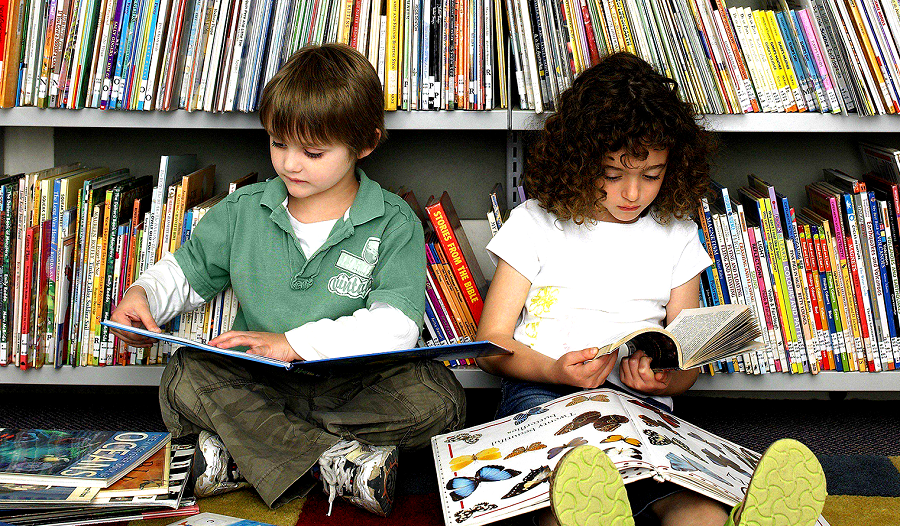 Picture of two kids reading in the library surrounded by books.
