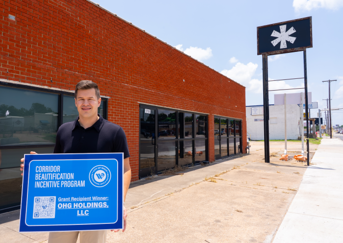 A man in front of a vacant brick building holding award recipient sign