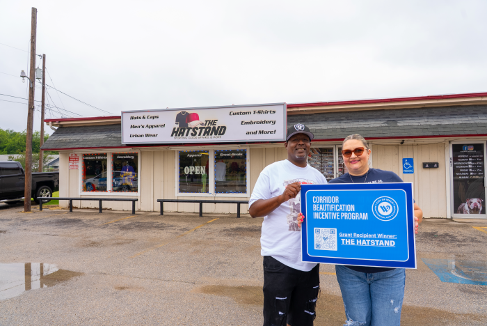 A man and woman in front of The Hatstand storefront holding award recipient sign