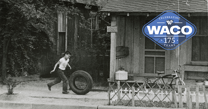 Black and white photo of a young boy playing with an old tire in front of a house