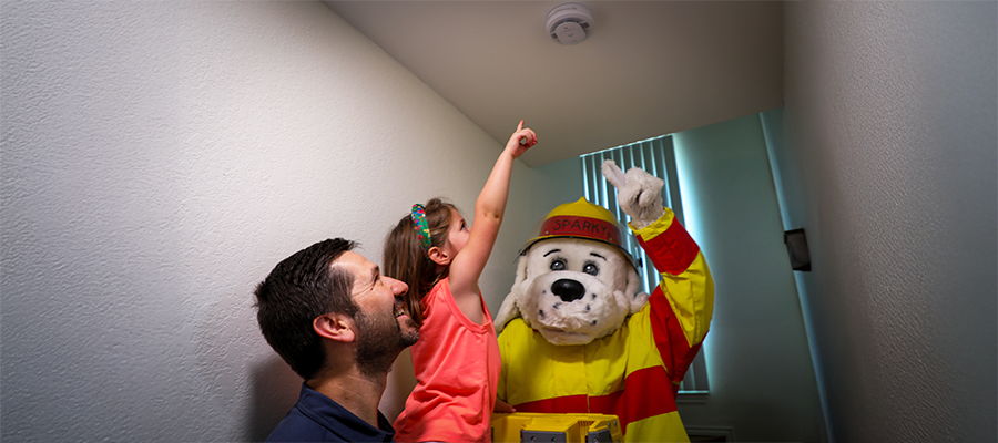 A dad, a girl, and Sparky the fire dog pointing at a smoke alarm on a ceiling